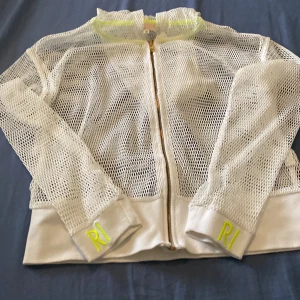 Vit meshjacka från River Island - Snygg vit meshjacka från River Island RI Active med dragkedja och neongula detaljer på muddarna. Jackan har långärmade ärmar och sportig känsla, perfekt för en cool och avslappnad stil. RI-logga broderad på båda ärmsluten. inte bara det utan under en varm sommar dag där det inte behövs jacka kan man ta fram en luftig kofta som är bekväm. 🥵☀️