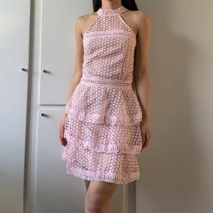 Ljusrosa spetsklänning med volanger - Supersöt ljusrosa kortklänning med halterneck och flera lager volanger. Klänningen har ett spetsigt, virkat mönster och är ärmlös. Dragkedja i ryggen.