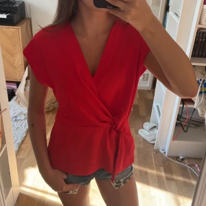 Röd omlottopp med knyt i midjan - Säljer en snygg röd omlottopp med knyt i midjan och v-ringning. Toppen har korta ärmar och en avslappnad passform som ger en cool och trendig look. Perfekt att matcha med jeansshorts eller kjol för en färgstark outfit.