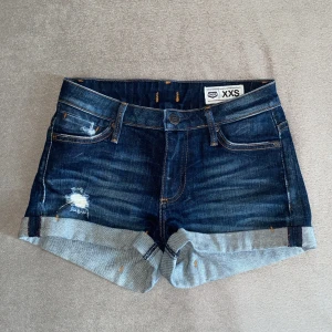 Mörkblå jeansshorts från Crocker - Snygga mörkblå jeansshorts från Crocker med upprullade benslut och slitna detaljer framtill. Klassisk femficksmodell med orangea sömmar och läderpatch bak i midjan. Perfekta för sommaren🤪🌸