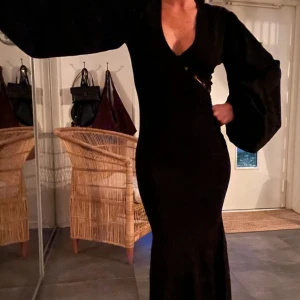 Adoore långklänning - Säljer en svart långklänning med djup v-ringning och dekorativa knappar framtill. Klänningen har långa ballongärmar och figurnära passform som framhäver siluetten. Perfekt för dig som vill ha en elegant och stilren look.