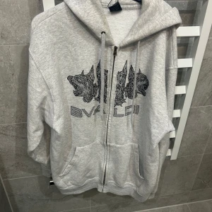 Grå hoodie med dragkedja från Bvaldi - Säljer en grå hoodie med dragkedja från Bvaldi. Tröjan har svart tryck framtill, huva med dragsko och två fickor. Perfekt för en avslappnad stil.