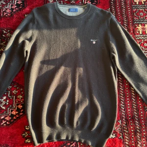 Mörkgrå stickad tröja från GANT - Säljer en mörkgrå stickad tröja från GANT med rund halsringning och diskret logga broderad på bröstet. Tröjan har långa ärmar och ribbade muddar vid ärmslut och nederkant. Storlek L aldrig använd!