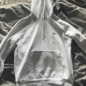 Vit hoodie från Lacoste - Säljer en klassisk vit hoodie från Lacoste med broderad logga på bröstet. Tröjan har huva med dragsko och en stor magficka framtill. Perfekt för en clean och stilren look. Inte en synlig fläck på tröjan då den typ är helt ny.