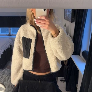 Vit teddyjacka med svart ficka - Säljer en vit teddyjacka med svart dragkedja och en stor svart ficka på bröstet. Jackan är croppad och har lång ärm. Perfekt för dig som vill ha en mysig och trendig look.