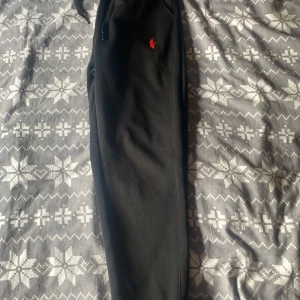Svarta mjukisbyxor från Polo Ralph Lauren - Svarta mjukisbyxor från Polo Ralph Lauren med röd broderad logga på ena benet. Byxorna har dragsko i midjan och muddar vid bensluten. Perfekta för en avslappnad stil och riktigt sköna att ha på sig. 