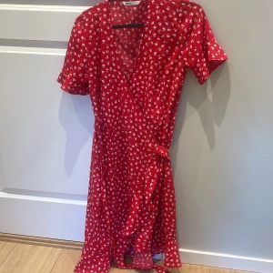 Röd omlottklänning med blommigt mönster - Superfin röd omlottklänning från Only med vitt blommönster. Klänningen har korta ärmar, knyt i midjan och v-ringad hals. Perfekt för sommarens alla tillfällen!