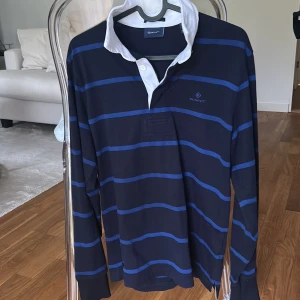 Blå randig pikétröja från Gant - Säljer en klassisk långärmad pikétröja från Gant i mörkblått med blå ränder och vit krage. Tröjan har knäppning vid halsen och är perfekt för en stilren look. Passar dig som gillar sportig och tidlös stil.