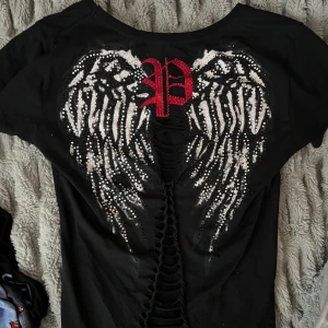 Svart t-shirt med vingar och snörning från Philipp Plein - Tar emot Bud! Högsta bud just nu är 250kr .Skambud undanbes! Svart t-shirt med v-ringning från Philipp Plein. Baksidan har stora  vingar och ett rött P i glitter. Snygg snörningsdetalj längs ryggen. Framsidan har ett mindre rött P i paljetter på bröstet. Äkta och vintage från min mamma som jag fått ärva . Storlek XL men passar S med en fin passform. 