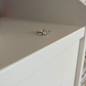 Silverring med hjärtformad sten från Pandora - Säljer en söt silverring från Pandora med en hjärtformad klar sten i mitten och små gnistrande stenar runt om. Ringen har en detaljerad, glittrig design och är perfekt för dig som gillar romantiska smycken. Perfekt som present till någon. Storlek 48 den minsta storleken. 