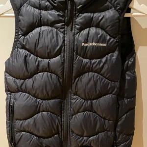 Svart dunväst från Peak Performance - Säljer en svart dunväst från Peak Performance med quiltad design och dragkedja framtill. Västen har en sportig look och är perfekt för lager-på-lager. Klassisk logga på bröstet och praktiska fickor med dragkedja.
