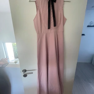Ljusrosa ärmlös långklänning med knappar och rosett - Säljer en ljusrosa långklänning med plisserad överdel, dekorativa knappar och svart rosett framtill. Klänningen är ärmlös och har fina spetsdetaljer runt bröstpartiet. Perfekt för sommarens tillställningar!