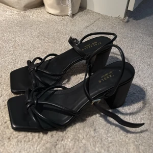 Svarta sandaler med klack från Stockholm Design Group - Säljer ett par svarta sandaler med hög blockklack och fyrkantig tå. ENDAST använda 1 gång! Skorna har tunna remmar med knyt runt vristen och stilren design. Perfekta för dig som gillar trendiga och eleganta skor. 