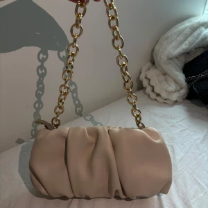 Beige axelväska med kedja - Beige axelväska med mjukt, rynkat material och en snygg guldfärgad kedja som axelrem. Väskan har dragkedja upptill.