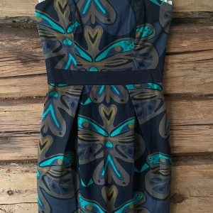 Svart klänning med fjärilsmönster - Säljer en svart klänning med stort grafiskt fjärilsmönster i blått, turkos, beige och gult. Klänningen har en markerad midja och är ärmlös med en figurnära passform. Perfekt för dig som vill sticka ut med ett unikt mönster.