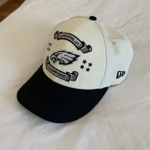 Philadelphia Eagles Super Bowl LII keps - Vit och svart keps från New Era med Philadelphia Eagles-logga och Super Bowl LII Champions brodyr framtill. NFL-märke baktill och Super Bowl LII-logga på sidan. Justerbar passform med snapback.