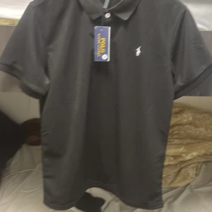 Svart pikétröja från Polo Ralph Lauren - Klassisk svart pikétröja från Polo Ralph Lauren med vit broderad logga på bröstet. Tröjan har krage, knappar framtill och är tillverkad i bomull. Perfekt för en stilren och tidlös look.