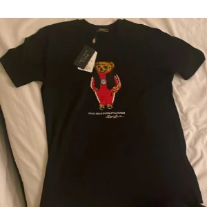 Svart t-shirt med björntryck från Ralph Lauren - Svart t-shirt från Ralph Lauren med ett färgglatt tryck av en björn på bröstet. Klassisk rund hals och korta ärmar. Perfekt för dig som gillar streetstyle och vill ha något unikt i garderoben.