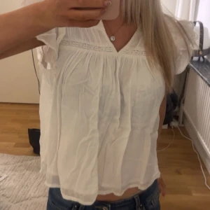 Vit blus med spetsdetaljer - Säljer en vit blus med v-ringning med spetsdetaljer vid axlar och bröst. Blusen har korta volangärmar. Ser skrynklig ut men går att stryka. Inga defekter, använd fåtal gånger. Den är i storlek 146/152 från gina young.🤗