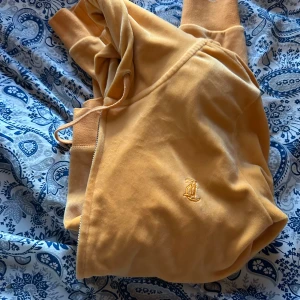 Gul hoodie från Juicy Couture - Säljer en mjuk och lyxig gul hoodie från Juicy Couture i velour med broderad JC-logga på bröstet. Tröjan har huva med dragsko och ribbade muddar. Perfekt för dig som gillar bekväma och snygga plagg!