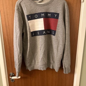 Grå sweatshirt från Tommy Jeans - Säljer en klassisk grå sweatshirt från Tommy Jeans med stort tryck på bröstet i vitt, rött och marinblått. Tröjan har rund halsringning och långa ärmar. Perfekt för en avslappnad och stilren look.