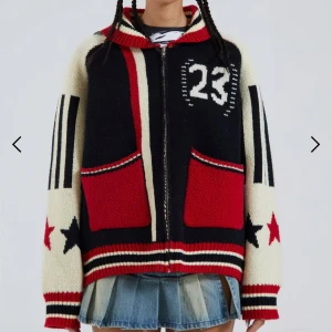 Jacka från Jaded London - Säljer en stickad jacka i svart, rött och vitt med dragkedja framtill. Jackan har röda fickor, randiga och stjärnmönstrade ärmar samt nummer 23 på bröstet. 