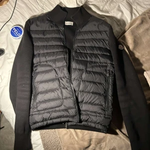 Svart quiltad jacka från Moncler - Säljer en svart quiltad jacka från Moncler med ribbad krage och ärmslut. Jackan har dragkedja framtill och två sidofickor med dragkedja. Klassisk Moncler-logga på ärmen och stilren design.
