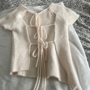 Beige fluffig topp med knytning - Supermjuk och fluffig beige topp med korta ärmar och unik knytning bak. Toppen har en lätt croppad passform och är tillverkad i ett syntetmaterial som ger en mysig känsla. Perfekt för dig som vill ha något annorlunda och trendigt.