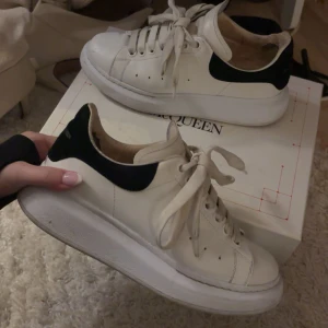 Vita sneakers från Alexander McQueen - Säljer ett par klassiska vita sneakers från Alexander McQueen med svart hälparti och tjock vit sula. Skorna har snörning och rund tå, samt diskret logga på hälen. Perfekta för dig som gillar stilrena och trendiga sneakers. De är använda men fortfarande i gott skick, de är lite slits i hälen där av priset!