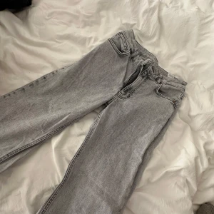 Gråa raka jeans - Säljer ett par ljusgrå jeans med rak passform och klassisk femficksdesign. Jeansen har dragkedja och knapp i midjan samt bälteshällor. Perfekta till vardags för en avslappnad stil.