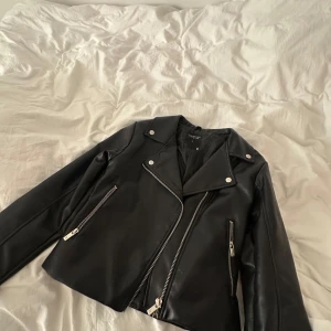 Svart skinnjacka med dragkedjor - Säljer en svart skinnjacka från Bik Bok med silvriga dragkedjor och knappar. Jackan har klassisk bikerstil med bred krage och två snedställda fickor framtill. Perfekt för en cool och stilren look.