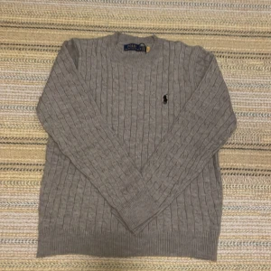 Ralph lauren ull tröja  - Säljer en klassisk grå kabelstickad tröja från Polo Ralph Lauren med broderad logga på bröstet. Tröjan har rund halsringning och långa ärmar. Perfekt för lager-på-lager under kyliga dagar.
