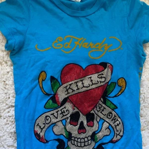 Blå Ed Hardy t-shirt med glittrigt tryck - Cool blå t-shirt från Ed Hardy med stort glittrigt tryck framtill av ett hjärta, dödskalle och texten 'Love Kills Slowly'. T-shirten har korta ärmar och klassisk passform. Perfekt för dig som gillar statement-plagg och färg!