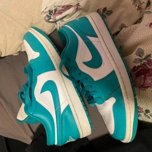 Nike Air Jordan 1 Low i turkos och vitt - Säljer ett par Nike Air Jordan 1 Low i turkos och vitt. (Ena plast grejen på skosnöret har gått av men märks inte av) Skorna har klassisk låg siluett, vit bas med turkosa paneler och snörning. Ikonisk Jumpman-logga på plösen och sidoswoosh i vitt. Perfekta för dig som gillar streetwear och sneakers.