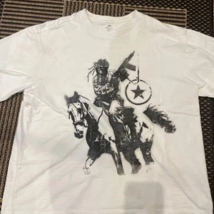 Vit t-shirt med tryck från H&M - As fet t-shirt från H&M med relaxed fit och ett stort svart tryck av en cowboy på häst med gevär och sköld med stjärna. Tröjan har några fläckar som igentigen inte är märkbar alls när man har på sig den. T-shirten har rund hals och korta ärmar, tillverkad i bomull. Tröjan köptes några år sen och finns ej att köpa i H&Ms butik mer, skriv om du har frågor 💕💕