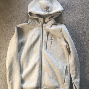 Ljusgrå hoodie från Sail Racing - Säljer en ljusgrå hoodie från Sail Racing med dragkedja framtill och en bröstficka med dragkedja. Jackan har huva med broderad logga och svart Sail Racing-tryck på bröstet. Perfekt för en sportig och avslappnad stil.