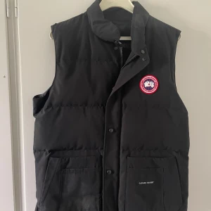 Svart dunväst från Canada Goose - Säljer en svart dunväst från Canada Goose med klassisk logga på bröstet och hög krage. Västen har tryckknappar och dragkedja framtill samt två stora fickor. Perfekt för lager-på-lager under kyliga dagar.