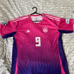 Tysklands fotbollströja rosa/lila - Säljer en riktigt snygg Tyskland fotbollströja från Adidas i rosa och lila med nummer 9 på bröstet. Tröjan har korta ärmar, vita detaljer och fyra stjärnor ovanför landslagsemblemet. Perfekt för dig som vill sticka ut på matchen! FÜLLKRUG 