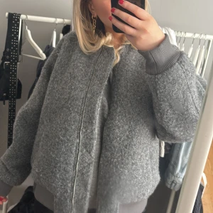 Grå bomberjacka med dragkedja - Säljer en snygg grå bomberjacka med dragkedja framtill. Jackan har en fluffig textur och ribbade muddar vid ärmslut och nederkant. Perfekt för kyligare dagar!