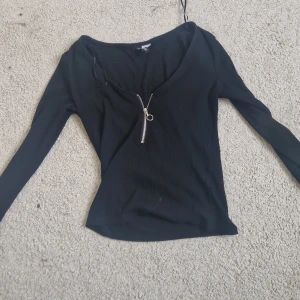 Svart ribbad långärmad topp med dragkedja - Svart långärmad topp med ribbad struktur och snygg v-ringning. Dragkedja framtill som detalj. Perfekt till jeans eller kjol för en stilren look.