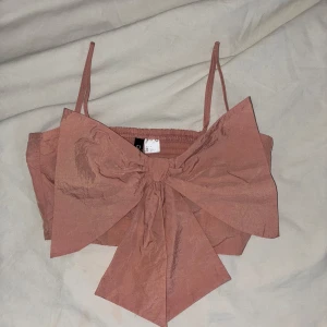 topp med rosett - Rosa croppad topp med smala axelband och stor rosett framtill. Prislappen är halv då det var en present som tyvärr aldrig kommit till användning