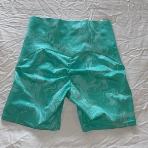 Turkosa träningstights-shorts från Gymshark - Säljer ett par turkosa träningstights-shorts från Gymshark i storlek XS. De har ett snyggt marmorliknande mönster och hög midja. Materialet är stretchigt och består av återvunnen polyester och elastan. Bara använd fåtal gånger, så väldigt fint skick!
