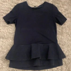 Mörkblå topp med volang - Säljer en mörkblå topp med korta ärmar och volang nertill. Toppen har en rund halsringning och är enkel men ändå stilren, perfekt att matcha med jeans eller kjol.