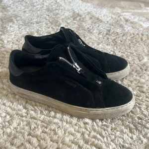 Svarta sneakers från Axel Arigato med dragkedja - Svarta sneakers från Axel Arigato i mocka med dragkedja framtill istället för klassisk snörning. Skorna har vit platt sula och diskret logga på sidan. Perfekta för en stilren och modern look.