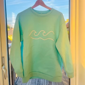 Turkis sweatshirt  - Säljer en turkos sweatshirt med våg-motiv framtill och tryck i nacken. Tröjan har rund hals och långa ärmar, perfekt för i sommar