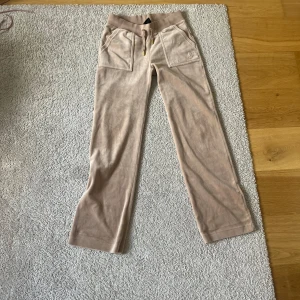 Beiga mjukisbyxor från Juicy Couture - Säljer ett par beiga mjukisbyxor från Juicy Couture med bred resår i midjan och snörning med gulddetaljer. Byxorna har raka ben och stora fickor framtill. Perfekta för en avslappnad stil.