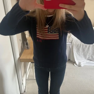 Mörkblå stickad tröja från Ralph Lauren - Säljer denna populära mörkblå stickade tröja från Ralph Lauren med amerikansk flagga och RL-broderi på bröstet. Tröjan har rund halsringning och långa ärmar med ribbade muddar. Perfekt för dig som gillar klassisk och tidlös stil. Nypris: 3000 kr.