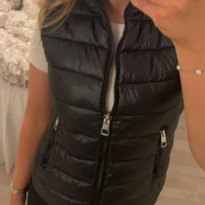 Svart dunväst med dragkedja från Mango - Snygg svart dunväst med dragkedja framtill och två fickor med dragkedja. Perfekt att slänga över en t-shirt för en sportig och trendig look. Västen har en lätt glansig yta och quiltad design.