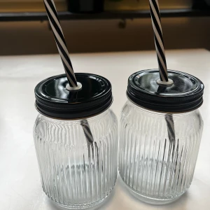  glasburk med svart lock och sugrör - Säljer en stilren glasburk med räfflad design, svart metallock och svartvitt randigt sugrör. Perfekt för kalla drycker och smoothies. Burken är genomskinlig och har en modern look.