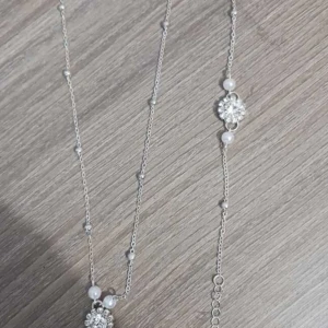 Silverfärgat halsband med pärlor och blomdetaljer - Elegant halsband i silverfärg med små vita pärlor och glittrande blomformade detaljer. Perfekt accessoar för att lyfta din outfit med en feminin touch. Justerbar längd för bästa passform.